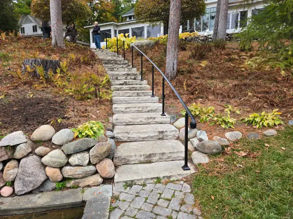 Black steel handrail on a long run of stone steps through a hillside garden toward a lake home